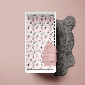 May include: A white crib with a floral patterned sheet and a pink blanket. A gray, fluffy rug is next to the crib. The sheet has a pattern of pink and blue flowers on a cream background.