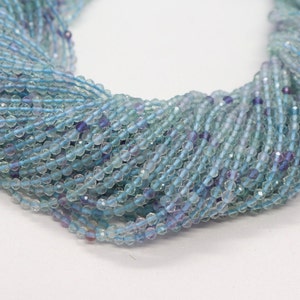 May include: A strand of light blue and purple faceted gemstone beads. The beads are small and round, and they are strung together on a clear thread.