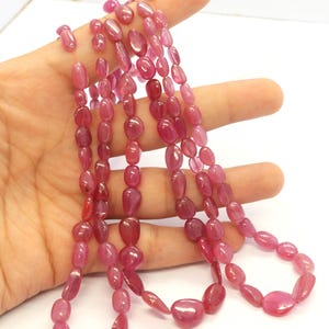 May include: A strand of polished, translucent pinkish-red gemstone beads with an irregular, rounded pebble shape. The beads are strung together in a continuous line, displaying varying sizes and a smooth, glossy surface texture throughout the strand.