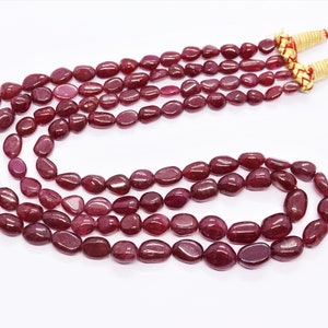 May include: A strand of red ruby gemstone beads, strung on a cord with gold tassels. The beads are oval shaped and have a smooth, polished finish.