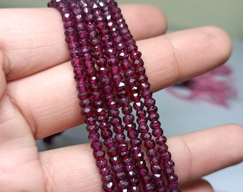 May include: A strand of deep red gemstone beads. The beads are faceted and have a slightly irregular shape. The strand is held in a hand, showing the beads in detail.