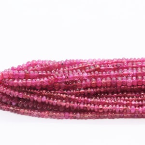 Può includere: Una collana di perle di rubino rosa, ogni perla è rotonda e leggermente allungata. Le perle sono infilate su un cordoncino rosso.