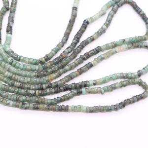 May include: Strands of small, square-cut emerald beads in varying shades of green and dark gray. The beads are strung together, creating a textured and organic look. The beads are displayed against a white background.