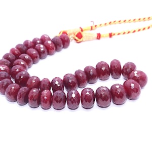 Puede incluir: Un collar o pulsera de cuentas facetadas de piedras preciosas de color rojo intenso. Las cuentas están ensartadas en un cordón dorado y rojo. Las piedras preciosas tienen una superficie pulida y reflectante, sobre fondo blanco.