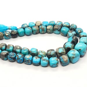 Puede incluir: Collar de cuentas de color turquesa con una mezcla de tonos azules y grises. Las cuentas tienen forma irregular y están ensartadas. Una borla azul brillante está unida al extremo del hilo. Las cuentas están sobre un fondo blanco.
