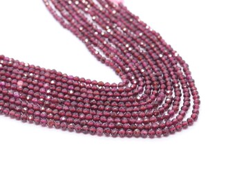 May include: Close-up of multiple strands of small, faceted, deep red garnet beads. The beads are strung together, creating a cascading effect against a white background. The beads are a rich, dark red color.
