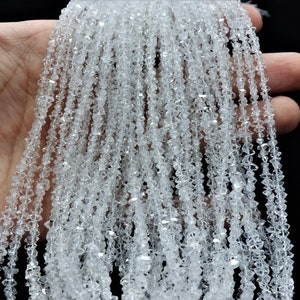 K&ouml;nnte beinhalten: Eine Str&auml;hne aus klaren, facettierten Kristallperlen. Die Perlen sind klein und haben eine diamantartige Form. Die Str&auml;hne wird in einer Hand gehalten, und die Perlen fallen herunter.