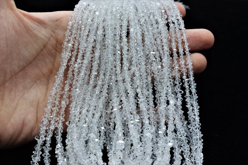 K&ouml;nnte beinhalten: Eine Str&auml;hne aus klaren, facettierten Kristallperlen. Die Perlen sind klein und haben eine diamantartige Form. Die Str&auml;hne wird in einer Hand gehalten, und die Perlen fallen herunter.