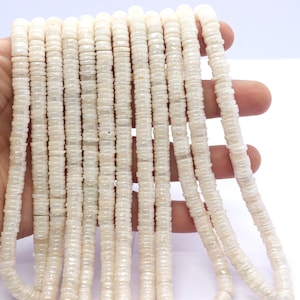 Puede incluir: Una hebra de cuentas de concha blanca iridiscente, cada cuenta es un cilindro con una parte superior e inferior ligeramente redondeadas. Las cuentas están enhebradas en un hilo transparente y tienen aproximadamente 10 mm de diámetro.