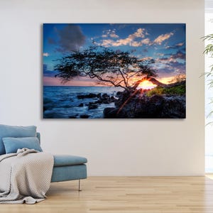May include: A canvas print of a coastal sunset scene. The image features a silhouetted tree on a rocky shore, with the sun setting behind it, casting a warm glow. The sky is a mix of blue, orange, and pink hues, with some clouds.
