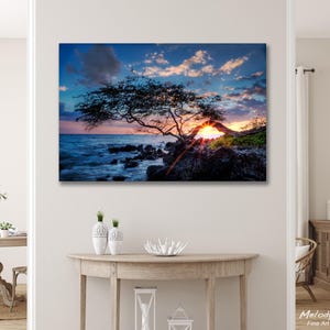 May include: A scenic canvas print of a sunset over the ocean. The image features a silhouetted tree with a vibrant sky of blues, oranges, and yellows. The ocean's waves crash against the rocky shore, creating a dramatic landscape.
