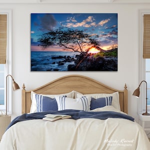 May include: A large framed canvas print of a sunset over the ocean. The image features a silhouetted tree on the rocky shore, with the sun setting behind it. The sky is a mix of blue, orange, and pink.
