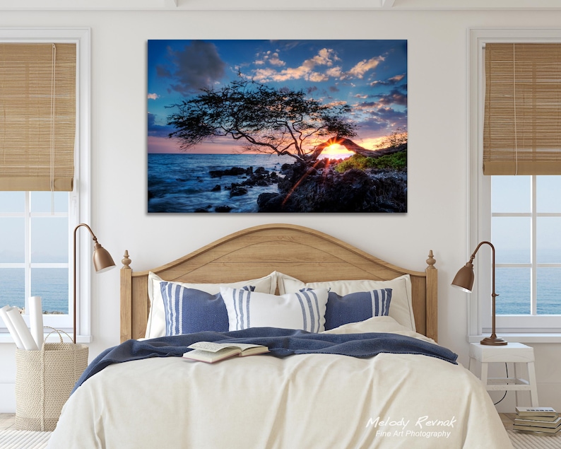 May include: A large framed canvas print of a sunset over the ocean. The image features a silhouetted tree on the rocky shore, with the sun setting behind it. The sky is a mix of blue, orange, and pink.