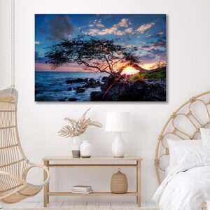 May include: A canvas print of a sunset over the ocean. A silhouetted tree frames the sun setting over the water. The sky is a mix of blue, orange, and pink. The print is displayed in a bedroom with a hanging chair and a wooden table.