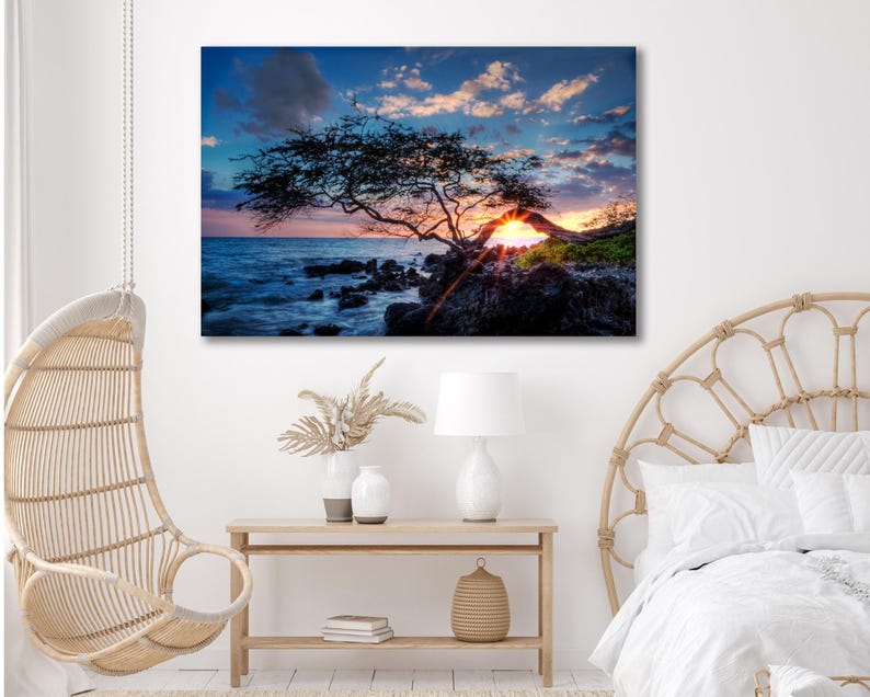 May include: A canvas print of a sunset over the ocean. A silhouetted tree frames the sun setting over the water. The sky is a mix of blue, orange, and pink. The print is displayed in a bedroom with a hanging chair and a wooden table.