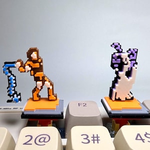 May include: Two 3D printed keycaps featuring pixelated characters. The first keycap is orange and blue and depicts a character holding a blue weapon. The second keycap is white and purple and depicts a character with a purple head. Both keycaps are on a keyboard with the keys F2 and F4 visible.