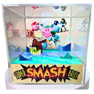 May include: A clear acrylic cube display case with a papercraft diorama inside. The diorama depicts characters from the video game Super Smash Bros. The characters are Donkey Kong, Link, and Kirby. The diorama is set against a blue and green background. The base of the display case has the words "Super Smash Bros" printed on it.