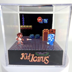 May include: A clear acrylic cube display case with a 3D diorama of a scene from the video game Kid Icarus. The diorama features the main character, Kid Icarus, fighting a blue monster. The diorama is set against a black background with the game's title, "Kid Icarus", printed in yellow and orange letters.