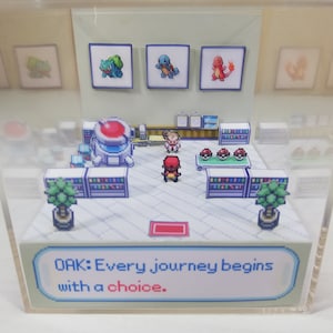 Puede incluir: Un diorama 3D de una escena de juego de Pokémon. La escena muestra a un entrenador Pokémon de pie frente al Profesor Oak, quien le está dando al entrenador una opción de Pokémon. El diorama está hecho de papel y cartón y se muestra en una caja de acrílico transparente. El texto en el diorama dice "OAK: Cada viaje comienza con una elección."
