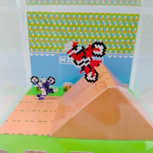 May include: A pixelated 8-bit video game character on a red motorcycle jumps over a ramp. The character is wearing a white helmet and is in mid-air. The ramp is brown and has a green base. The background is a blue sky with a white and yellow checkered pattern.
