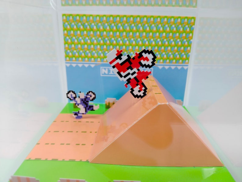 May include: A pixelated 8-bit video game character on a red motorcycle jumps over a ramp. The character is wearing a white helmet and is in mid-air. The ramp is brown and has a green base. The background is a blue sky with a white and yellow checkered pattern.