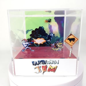 Pode incluir: Uma vitrine de acrílico transparente com um diorama de uma cena de videogame de Earthworm Jim. A cena apresenta o personagem Earthworm Jim disparando uma arma em uma criatura com um crânio de vaca. O diorama está situado em um fundo verde e roxo. A vitrine tem uma base branca com as palavras "Earthworm Jim" impressas nela.