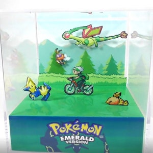 May include: A 3D diorama of the Pokemon Emerald Version game. The diorama features a green background with a forest scene. There are several Pokemon characters in the diorama, including a green and yellow flying Pokemon, a blue and yellow Pokemon, a yellow Pokemon, and a trainer riding a bike. The diorama also features the text "Pokemon Emerald Version".
