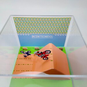May include: A clear acrylic display case with a miniature papercraft scene of a video game. The scene features two pixelated characters on dirt bikes, one jumping over a ramp. The background is a blue sky with the word "NINTENDO" printed in white.