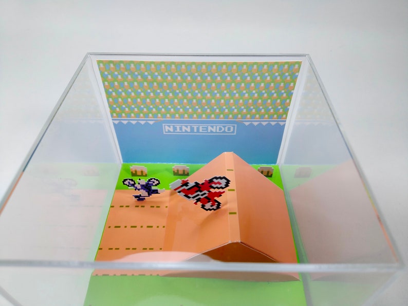 May include: A clear acrylic display case with a miniature papercraft scene of a video game. The scene features two pixelated characters on dirt bikes, one jumping over a ramp. The background is a blue sky with the word "NINTENDO" printed in white.