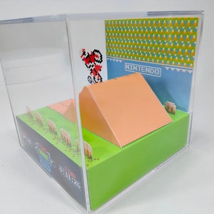 May include: A 3D papercraft diorama of the classic Nintendo video game, Excitebike. The diorama features a pixelated character riding a motorcycle over a ramp, with a green grassy background and a blue sky with the word "NINTENDO" printed on it.