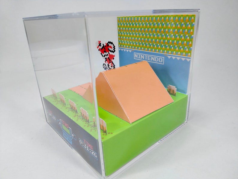 May include: A 3D papercraft diorama of the classic Nintendo video game, Excitebike. The diorama features a pixelated character riding a motorcycle over a ramp, with a green grassy background and a blue sky with the word "NINTENDO" printed on it.