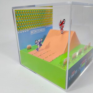 May include: A 3D diorama of the classic Nintendo video game, Excitebike. The diorama features a pixelated representation of the game's environment, including a dirt track, a ramp, and a finish line. The diorama also includes a pixelated representation of the game's main character, a biker, performing a stunt on the ramp. The diorama is enclosed in a clear acrylic case.