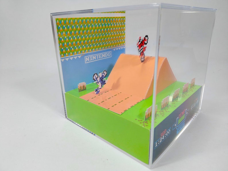 May include: A 3D diorama of the classic Nintendo video game, Excitebike. The diorama features a pixelated representation of the game's environment, including a dirt track, a ramp, and a finish line. The diorama also includes a pixelated representation of the game's main character, a biker, performing a stunt on the ramp. The diorama is enclosed in a clear acrylic case.