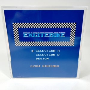May include: A blue Nintendo game cartridge with a checkered border and the text "EXCITEBIKE" in white with a blue outline. The text "SELECTION A", "SELECTION B", and "DESIGN" are printed in white below the title. The text "&copy;1984 NINTENDO" is printed in white at the bottom of the cartridge.