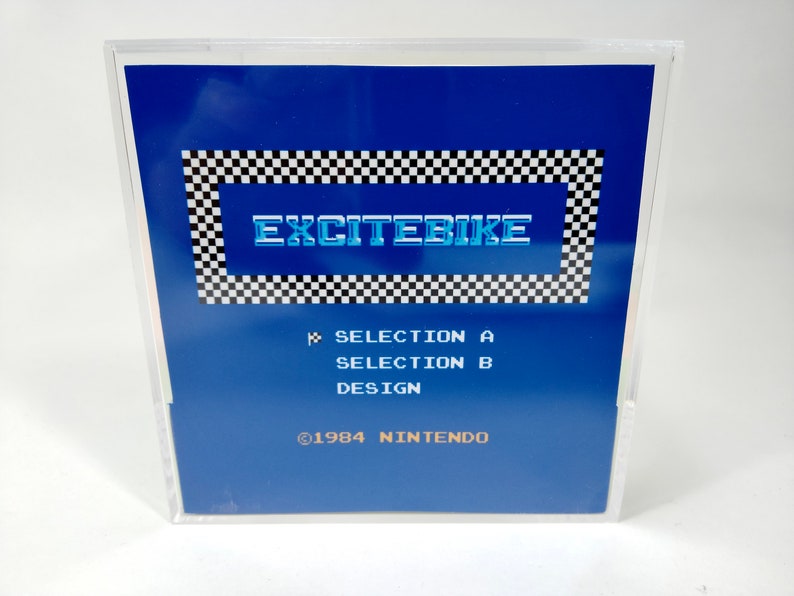 May include: A blue Nintendo game cartridge with a checkered border and the text "EXCITEBIKE" in white with a blue outline. The text "SELECTION A", "SELECTION B", and "DESIGN" are printed in white below the title. The text "&copy;1984 NINTENDO" is printed in white at the bottom of the cartridge.