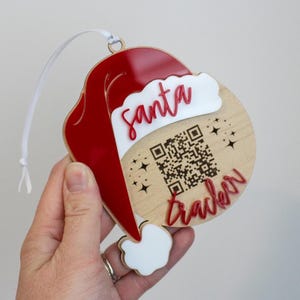 May include: A round wooden Christmas ornament shaped like a Santa hat with a red and white acrylic overlay. The ornament features the text "santa tracker" in red script and a central QR code surrounded by small engraved star icons and a white ribbon.