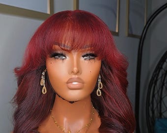 May include: A mannequin head displaying a burgundy wig with a fringe and wavy layers. The wig is styled with a side part and features a dark root. The mannequin is adorned with gold earrings and a layered gold necklace.