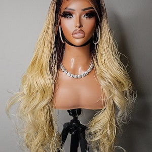 May include: A blonde and brown braided wig with a lace front on a wig stand. The wig is styled in loose waves.