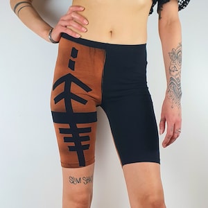 Op de afbeelding: Zwarte en bruine fietsbroek met een grafisch ontwerp op het linkerbeen. Het ontwerp bevat zwarte lijnen en de tekst "SUM SHA".