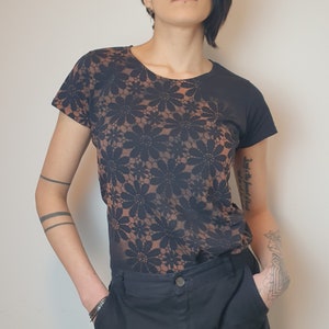 Op de afbeelding: Een persoon draagt een zwart shirt met korte mouwen met een bruin bloemenpatroon en een zwarte cargo broek.