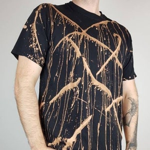 Könnte beinhalten: Ein schwarzes T-Shirt mit einem Design aus Bleichmittelspritzern. Das Design zeigt eine Reihe von braunen Linien und Tropfen.