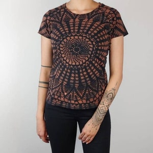 Op de afbeelding: Zwart T-shirt met korte mouwen en een bruin, cirkelvormig mandala-patroon. De top wordt gecombineerd met een zwarte skinny jeans en zwarte enkellaarsjes. De outfit wordt gepresenteerd tegen een witte muur en een houten vloer.