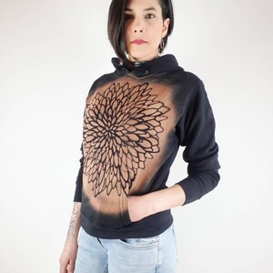Pu&ograve; includere: Una felpa con cappuccio nera con un design a mandala marrone e nero sul davanti. La felpa ha un cappuccio con coulisse e una tasca a marsupio.