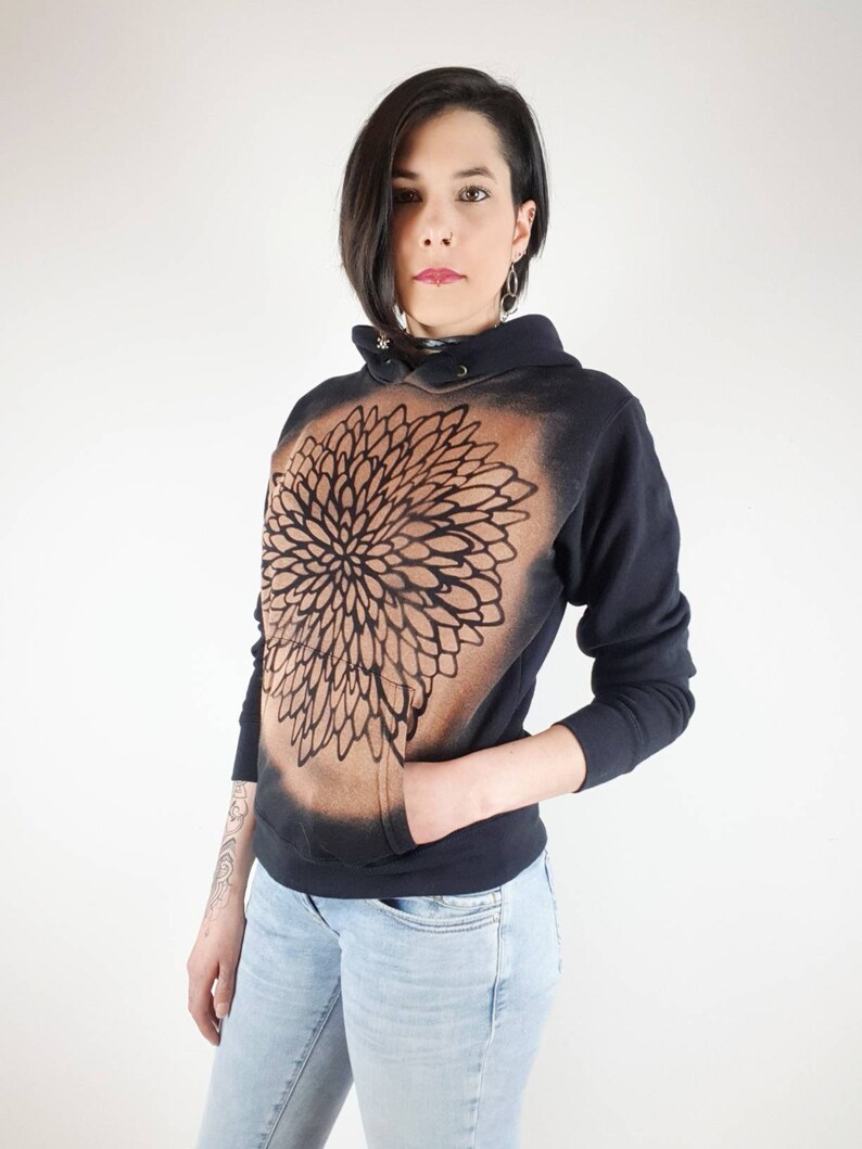 Pu&ograve; includere: Una felpa con cappuccio nera con un design a mandala marrone e nero sul davanti. La felpa ha un cappuccio con coulisse e una tasca a marsupio.