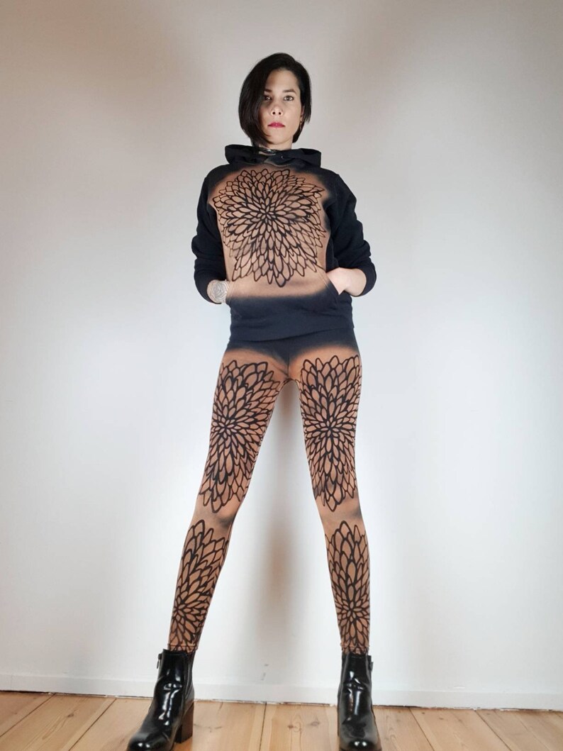 Pu&ograve; includere: Una felpa nera con un grande disegno di mandala sul davanti, abbinata a leggings neri e marroni con un disegno di mandala simile. La modella indossa stivaletti neri alla caviglia.