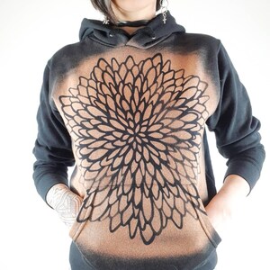 Pu&ograve; includere: Felpa con cappuccio nera con un design a mandala marrone e nero. La felpa ha un cappuccio con coulisse e una tasca a marsupio.