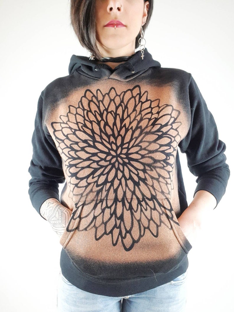 Pu&ograve; includere: Felpa con cappuccio nera con un design a mandala marrone e nero. La felpa ha un cappuccio con coulisse e una tasca a marsupio.