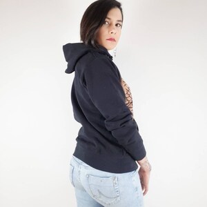 Pu&ograve; includere: Una donna indossa una felpa con cappuccio nera e jeans blu chiaro. Sta guardando oltre la sua spalla.