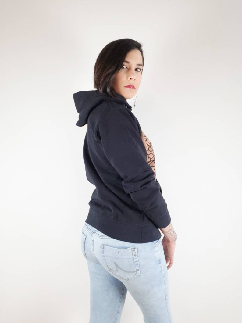 Pu&ograve; includere: Una donna indossa una felpa con cappuccio nera e jeans blu chiaro. Sta guardando oltre la sua spalla.