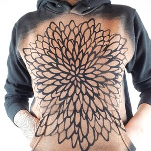 Pu&ograve; includere: Felpa con cappuccio nera con un grande disegno floreale marrone e nero sul davanti. Il disegno &egrave; un motivo a forma di mandala con una forma circolare.